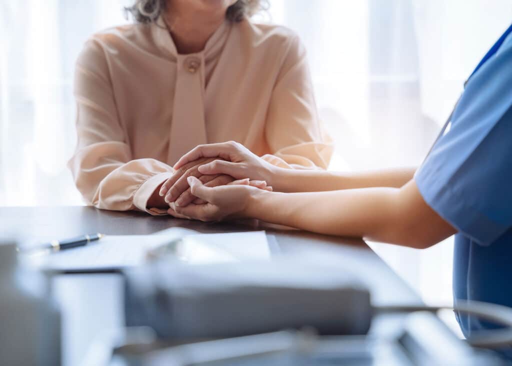 Discussing Patience Assistance Programs, A nurse holds an elderly woman's hands as they talk.