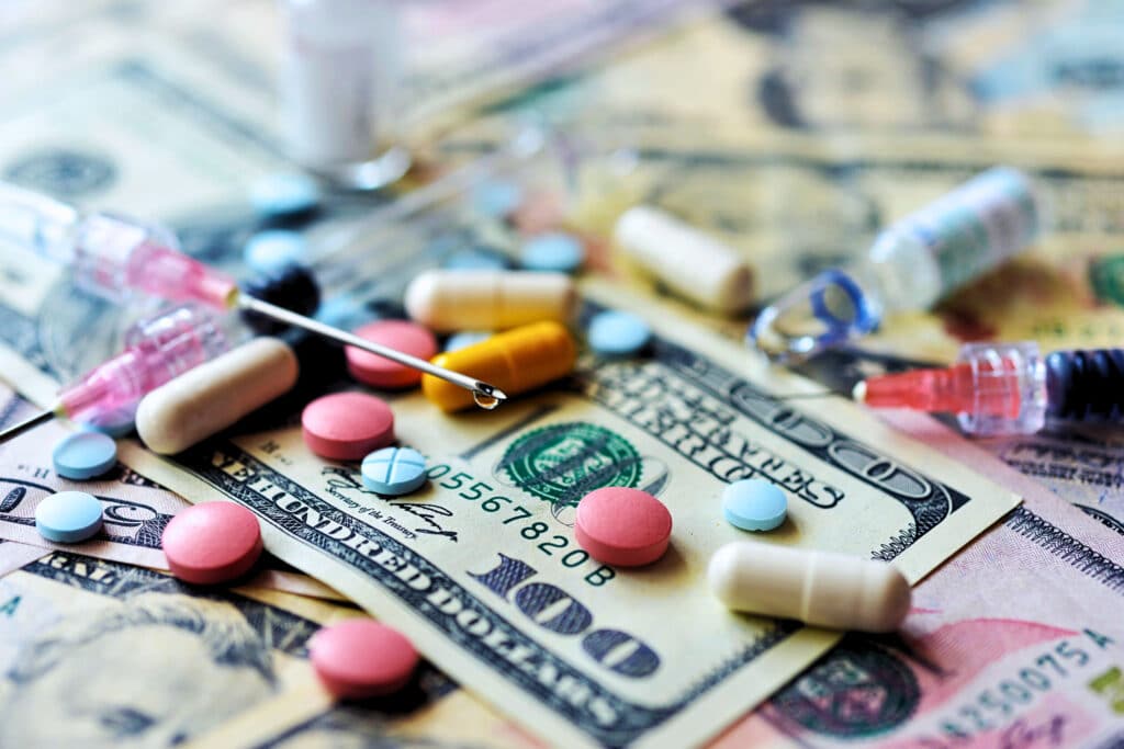 pills and insulin needles sit atop 0 bills, representing the pain of expensive prescription medications.